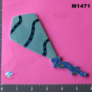 May include: A light blue ceramic decorative piece with dark blue wavy lines and black dots. The piece has a unique shape and a blue, yellow, and white decorative tail. The text "M1471" is visible.