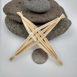May include: A handmade straw St. Brigid's cross, a traditional Irish craft, is displayed. The cross is made of woven straw and is positioned in front of two gray stones. A US quarter is placed near the cross for size comparison.