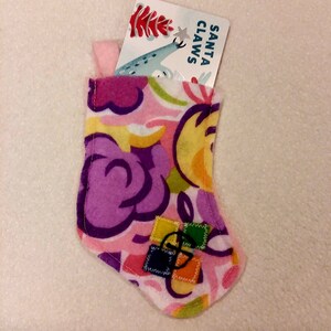 May include: A small, colorful Christmas stocking with a floral pattern in pink, purple, yellow, and white. The stocking contains a card that reads "SANTA CLAWS". The stocking has a small loop for hanging and is decorated with small, colorful patches.