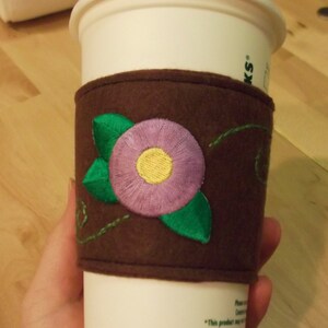 May include: A brown felt coffee cup sleeve featuring an embroidered purple flower with a yellow centre and green leaves. The sleeve is wrapped around a white disposable coffee cup with a white lid, displaying the Starbucks logo and text.