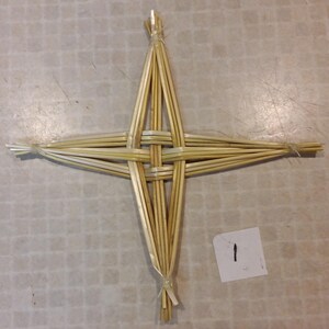 May include: A woven straw cross with a central square and four radiating arms. The natural-colored straw is bound together with twine at the center and the ends of the arms. A small white paper with the number "1" is in the lower right corner.