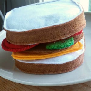 May include: A felt sandwich with brown bread, white filling, yellow cheese, a red tomato slice, and a green pickle. The sandwich is on a white plate, and the felt is stitched together.