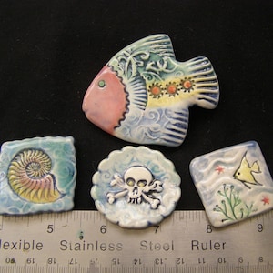 May include: A collection of ceramic art pieces featuring marine life and nautical themes. Includes a fish, a square with a nautilus, a round piece with a skull and crossbones, and a square with a fish and seaweed. The pieces are hand-painted with vibrant colors.