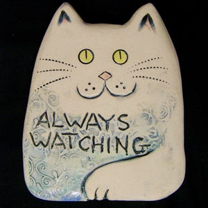 May include: A cream-colored ceramic cat figurine with yellow eyes and the text "ALWAYS WATCHING." The cat has a blue and white patterned design on its body and a smiling face. The cat has a black background.