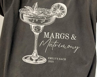 Może przedstawiać: Szara koszulka oversize z nadrukiem szklanki do margarity i napisem "MARGS & MATRIMONY" oraz "EMILY'S BACH 2025". Grafika jest biała. Koszulka ma krótkie rękawy.
