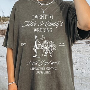 Op de afbeelding: Pepperkleurig T-shirt met witte tekst: "I went to Mike & Emily's Wedding & all I got was a hangover and this lousy shirt." Met een afbeelding van een champagnefles en bloemen. Het jaar 2025 staat op het shirt.
