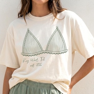 May include: Cream-coloured t-shirt with a green line drawing of a bra. The shirt has the text "Key West, FL est. 2026". The model is wearing green shorts. The t-shirt is a casual, comfortable style.