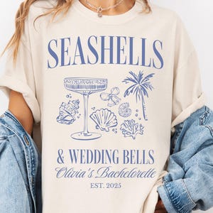 May include: Ivory t-shirt with a blue graphic print that says "Seashells & Wedding Bells Olivia's Bachelorette Est. 2025". The graphic includes a cocktail glass, seashells, and a palm tree.