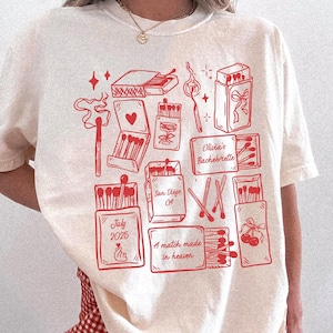 Könnte beinhalten: Cremefarbenes T-Shirt mit einem karmesinroten Aufdruck, der verschiedene Streichholzschachtel-Designs und den Text "Olivia's Bachelorette", "July 2025" und "A match made in heaven" zeigt. Das Shirt hat kurze Ärmel.