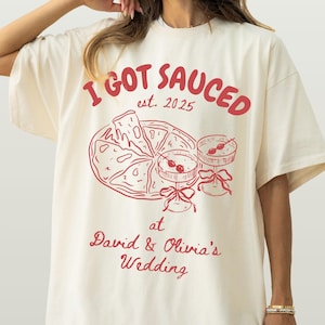 May include: Ivory t-shirt with a crimson print that says "I GOT SAUCED est. 2025" with a drawing of pizza slices and cocktails. The text below the graphic says "at David & Olivia's Wedding".