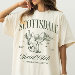 May include: Ivory-colored t-shirt with a moss green graphic. The graphic includes the word "SCOTTSDALE" above a desert scene with a cactus, cocktail, and mountains. Below the scene is "Social Club" and "OLIVIA'S BACHELORETTE".