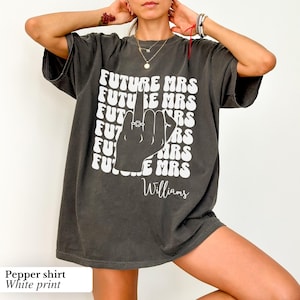 May include: A dark gray oversized t-shirt with a white print of a hand giving the middle finger with a ring on it. The text "FUTURE MRS" is repeated in a retro font and the name "Williams" is printed below the hand.