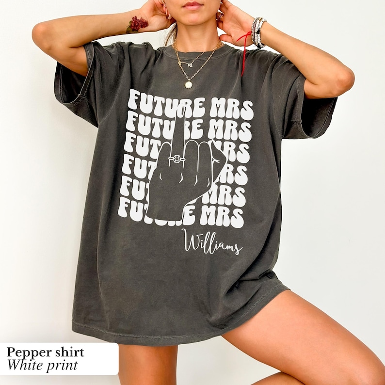 May include: A dark gray oversized t-shirt with a white print of a hand giving the middle finger with a ring on it. The text "FUTURE MRS" is repeated in a retro font and the name "Williams" is printed below the hand.