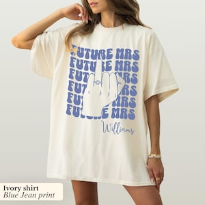May include: Ivory t-shirt with a blue jean print of a hand with a ring on the middle finger, and the text "FUTURE MRS" repeated multiple times. The text "Williams" is printed below the hand.