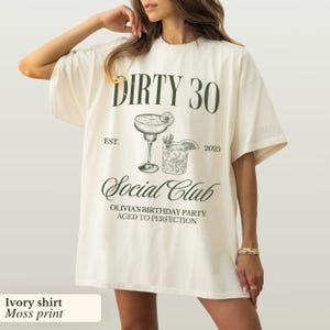 May include: Ivory t-shirt with a moss green print that says "Dirty 30 Est. 2025 Social Club Olivia's Birthday Party Aged to Perfection". The print also includes a drawing of two cocktails.