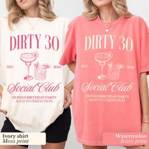 May include: Two t-shirts with the text "Dirty 30 Social Club" in white with a pink outline. The text "Olivia's Birthday Party Aged to Perfection" is below the main text. The shirts are in ivory and watermelon colors. The ivory shirt has a moss print and the watermelon shirt has an ivory print.