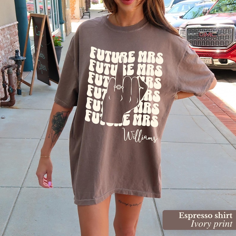 May include: Espresso brown t-shirt with a white print of a hand with a ring on the finger and the text "FUTURE MRS" repeated multiple times. The text "Williams" is printed below the hand.