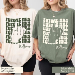 May include: Two t-shirts with a graphic print of a hand with a ring on the middle finger. The text on the shirts reads "FUTURE MRS" repeated multiple times. The shirts are in ivory and moss green colors. The text on the shirts is in a dark green color. The text "Williams" is printed at the bottom of the shirts.