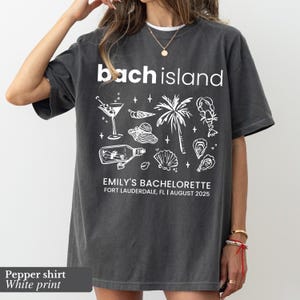 May include: Dark grey t-shirt with white graphic text and illustrations. The shirt reads "bach island" above a design of a cocktail, palm tree, and other beach-themed images. Below the design is "EMILY'S BACHELORETTE FORT LAUDERDALE, FL | AUGUST 2025".