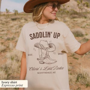 May include: Ivory t-shirt with a brown print of a cowboy boot and hat with the text "SADDLIN' UP EST. 2025 Olivia's Last Rodeo SCOTTSDALE, AZ"