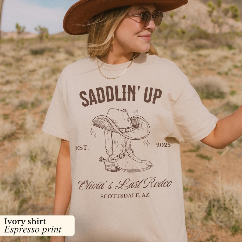 May include: Ivory t-shirt with a brown print of a cowboy boot and hat with the text "SADDLIN' UP EST. 2025 Olivia's Last Rodeo SCOTTSDALE, AZ"