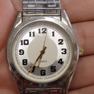 Pu&ograve; includere: Un orologio da polso argentato con quadrante bianco e numeri neri. L'orologio ha un cinturino in metallo argentato e dice "QUARTZ" sul quadrante.