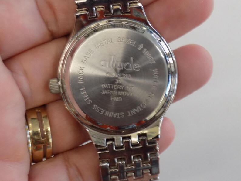 Puede incluir: Un reloj de pulsera de acero inoxidable plateado con un bisel de metal plateado y una correa plateada. La esfera del reloj est&aacute; marcada con la palabra "allude" y el texto "Steel Back Base Metal Bezel 100ft Water Resistant Stainless Steel".