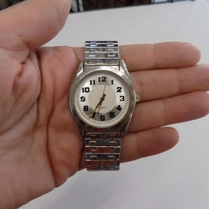Pu&ograve; includere: Un orologio da polso argentato con quadrante bianco e numeri neri. L'orologio ha un cinturino in metallo argentato.