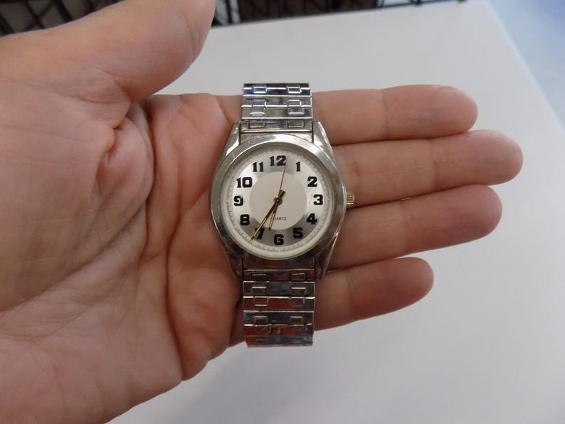 Pu&ograve; includere: Un orologio da polso argentato con quadrante bianco e numeri neri. L'orologio ha un cinturino in metallo argentato.