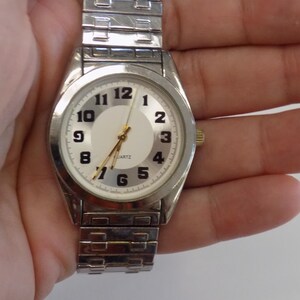 Pu&ograve; includere: Un orologio da polso argentato con quadrante bianco e numeri neri. L'orologio ha un cinturino in metallo argentato.