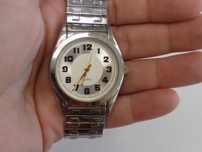 Pu&ograve; includere: Un orologio da polso argentato con quadrante bianco e numeri neri. L'orologio ha un cinturino in metallo argentato.