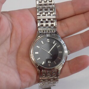 Puede incluir: Un reloj plateado con una esfera negra y una correa plateada. La esfera del reloj tiene un diamante en la posici&oacute;n de las 12 en punto y la palabra "DIAMOND" impresa debajo.