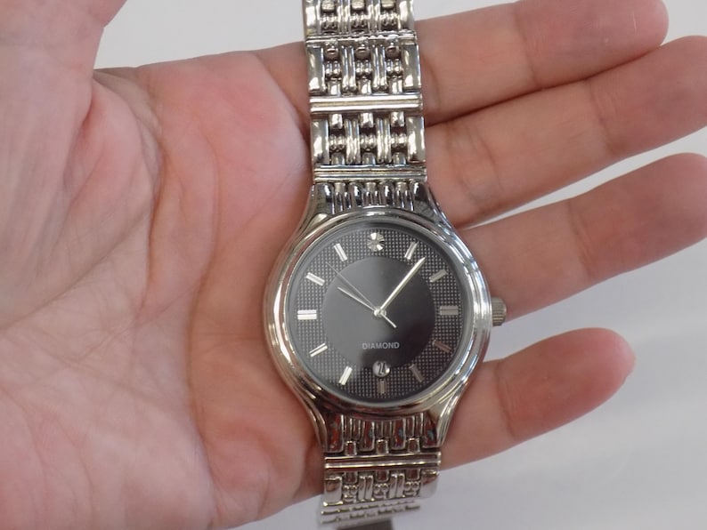 Puede incluir: Un reloj plateado con una esfera negra y una correa plateada. La esfera del reloj tiene un diamante en la posici&oacute;n de las 12 en punto y la palabra "DIAMOND" impresa debajo.