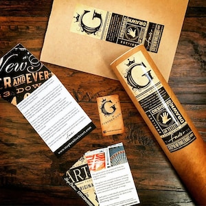 Puede incluir: Una colecci&oacute;n de impresiones art&iacute;sticas y materiales promocionales, incluyendo un p&oacute;ster enrollado y pegatinas, que muestran dise&ntilde;o gr&aacute;fico. Los art&iacute;culos presentan el logotipo y la marca del artista, con una paleta de colores marr&oacute;n, negro y blanco. El texto incluye "Original Artwork" y "Designed".