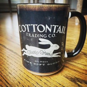 May include: A black ceramic mug with the text "COTTONTAIL TRADING CO." and a white rabbit graphic. The mug also features the text "TRADE MARK", "MFG. IN U.S.A.", "FINE SOFT GOODS", and "EST. 1872". The handle is black with white accents.
