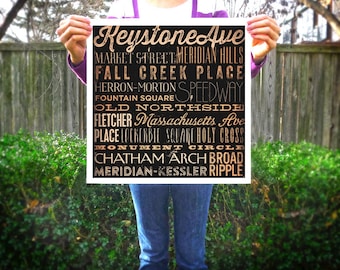 Peut inclure: Une affiche noire et blanche avec des lettres dorées indiquant des noms de rues tels que "Keystone Ave" et "Market Street". L'affiche est tenue devant un buisson vert et une clôture en bois.