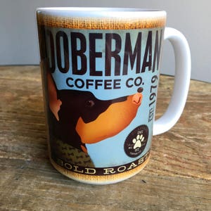 May include: A white ceramic coffee mug featuring a Doberman Pinscher illustration. The mug has the text "DOBERMAN COFFEE CO." and "BOLD ROAST" on a light blue background. The mug also has a paw print logo and the text "EST. 1979".