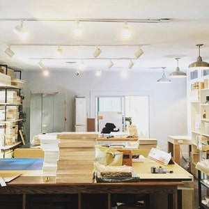 Puede incluir: Interior de un estudio de arte con mesas de trabajo, estanter&iacute;as e iluminaci&oacute;n de riel. Se ven pilas de paneles de madera y diversos materiales de arte. El espacio est&aacute; bien iluminado, lo que sugiere un espacio de trabajo creativo.