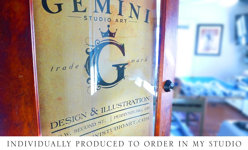 May include: A close-up of a wooden sign with a gold background and black text. The sign reads "GEMINI STUDIO ART" and features a stylized "G" with a crown above it. The text "trade mark" is also visible. The sign also includes the text "DESIGN & ILLUSTRATION", "W. SECOND ST. | PERRYSBURG, OH", and "NISTUDIOART.COM".