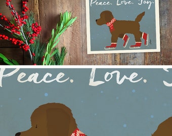 May include: A print featuring a brown dog wearing a red and white patterned scarf and ice skates. The text "Peace. Love. Joy." is at the top. The background is a muted blue with white snowflakes. The print is on a wooden background.