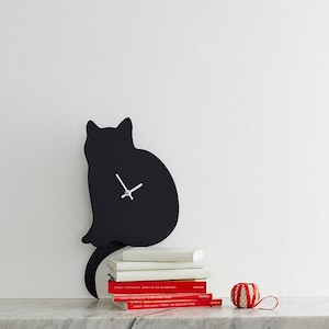 May include: Black cat silhouette wall clock with silver hands. The clock is hanging on a white wall above a stack of books with red spines. The books have the title "Ernest Hemingway" on the spines. A red and white striped ornament is on the shelf below the clock.