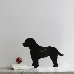 May include: Black silhouette of a dog with a clock face in the center. The dog is standing on a white surface with a red ball and books in front of it.