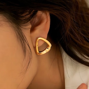 Puede incluir: Pendientes de oro en forma de triángulo con un centro hueco. Los pendientes son pequeños y delicados.