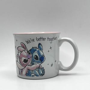 May include: A white ceramic mug with a pink interior and a curved handle. The mug features a colorful illustration of two cartoon characters embracing, with the text "We're better together".