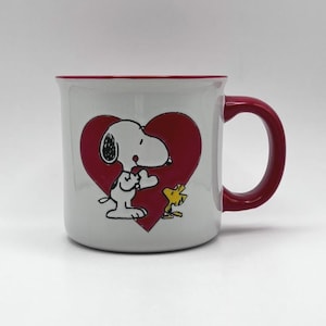 Pode incluir: Caneca de cerâmica branca com interior, alça e borda vermelhos. Apresenta um grande coração vermelho com Snoopy e um pássaro amarelo. Ideal para café ou chá.
