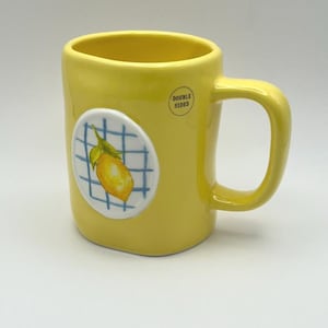 May include: Yellow ceramic mug with a rounded handle and a lemon illustration on a white and blue checkered background. The mug has the words "DOUBLE SIDED" printed on the side.