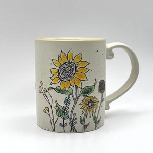 Pode incluir: Caneca de cerâmica com um design floral pintado à mão, apresentando um grande girassol amarelo e outras flores silvestres. A caneca tem uma base creme com uma textura salpicada e uma alça curva. As flores são delineadas em preto.