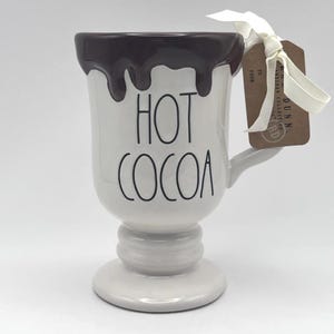 Peut inclure: Mug en céramique blanche avec une base en piédestal et une anse. Le bord est peint avec un motif dégoulinant marron foncé. Les mots "HOT COCOA" sont imprimés en noir. Une étiquette est attachée avec un ruban blanc.