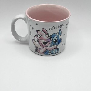 May include: White ceramic mug with a pink interior and rim. The mug features a colorful illustration of two cartoon characters embracing, with the text "We're better together." The mug has a curved handle.