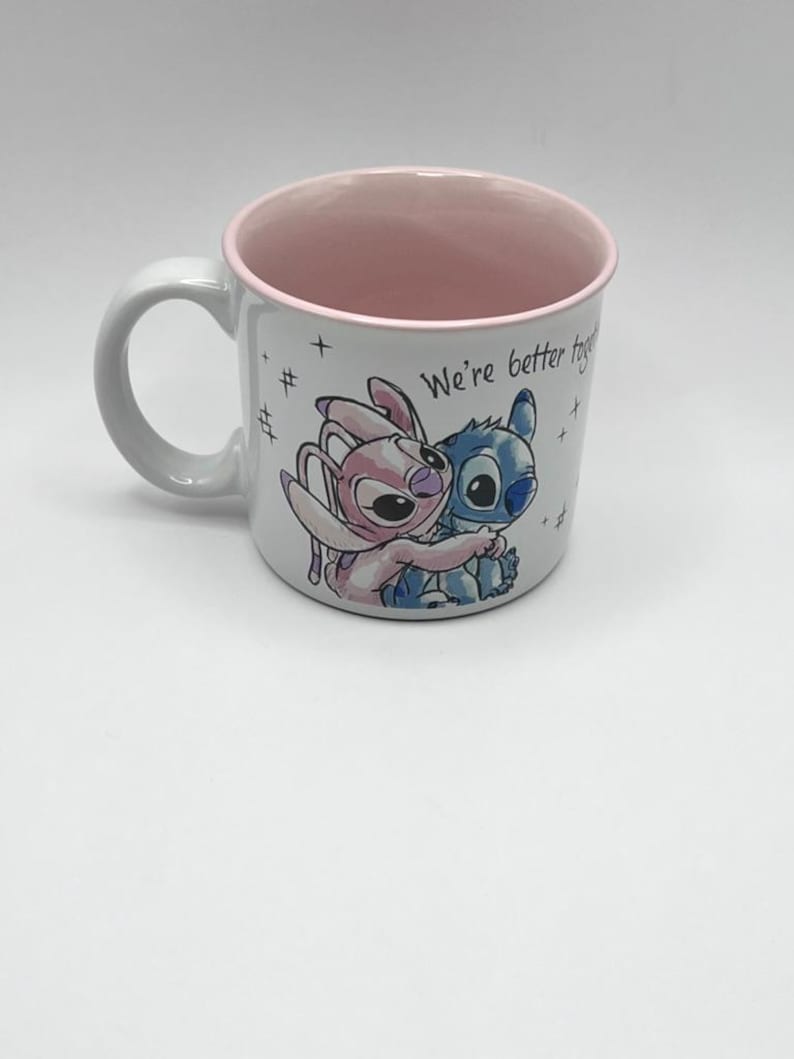 May include: White ceramic mug with a pink interior and rim. The mug features a colorful illustration of two cartoon characters embracing, with the text "We're better together." The mug has a curved handle.
